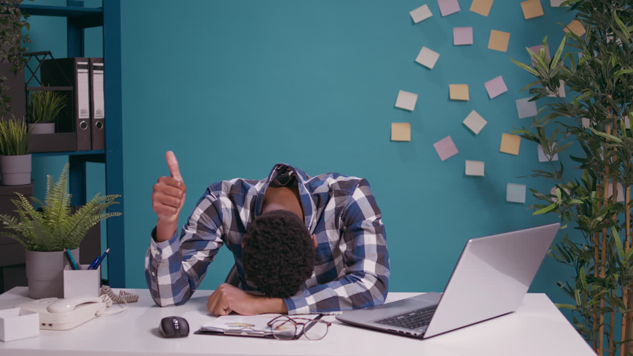 Exhausted worker putting head on desk and showing thumbs up sign