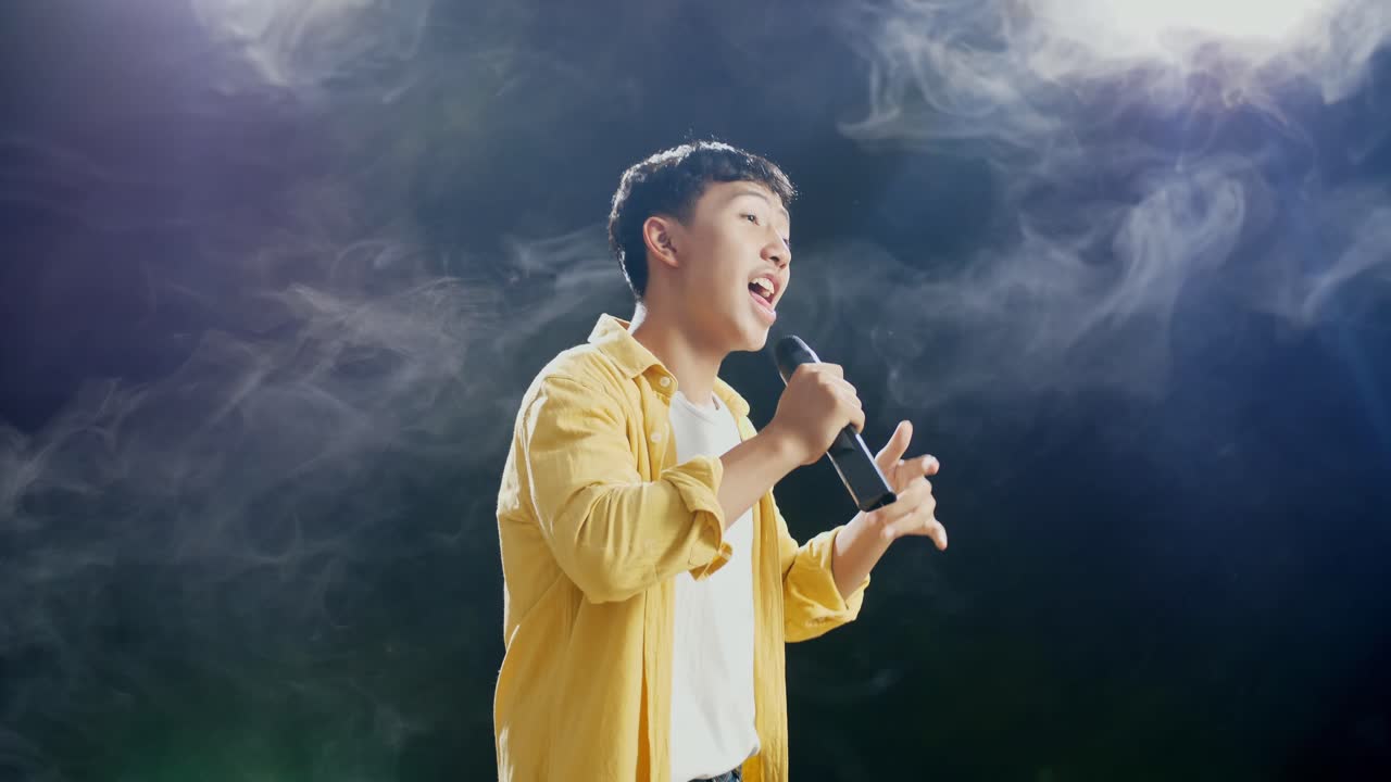 젊은 아시아 소년이 마이크를 들고 색 연기와 검은색 배경에서 춤추고 노래하는 측면 시각