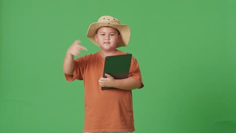 모자를 입은 아시아 관광 소년은 미소 짓고 녹색 화면 배경의 태블릿을 가리키고 있습니다. 소년 연구자는 여행, 관광, 모험 개념의 무언가를 조사합니다.