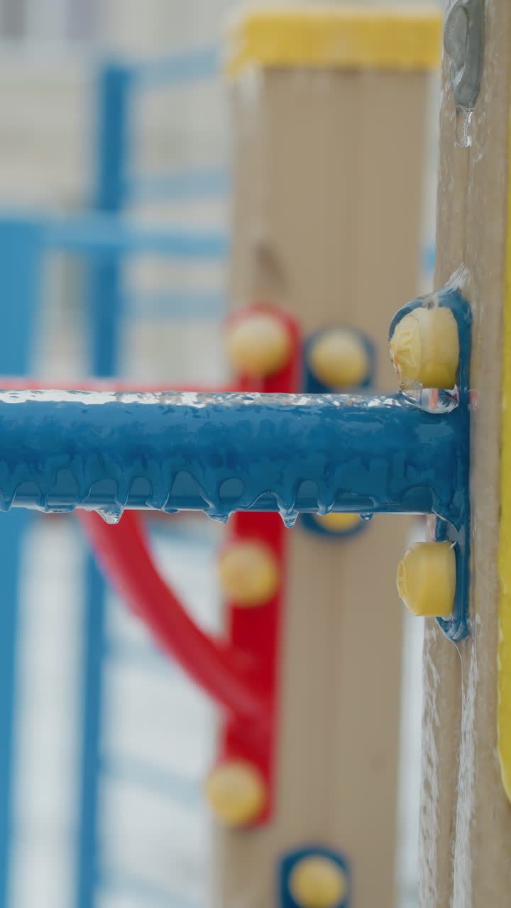Close-up de uma corda azul presa a uma superfície amarela coberta de gelo, destacando os detalhes delicados da geada de inverno e estruturas borradas de playground ao fundo