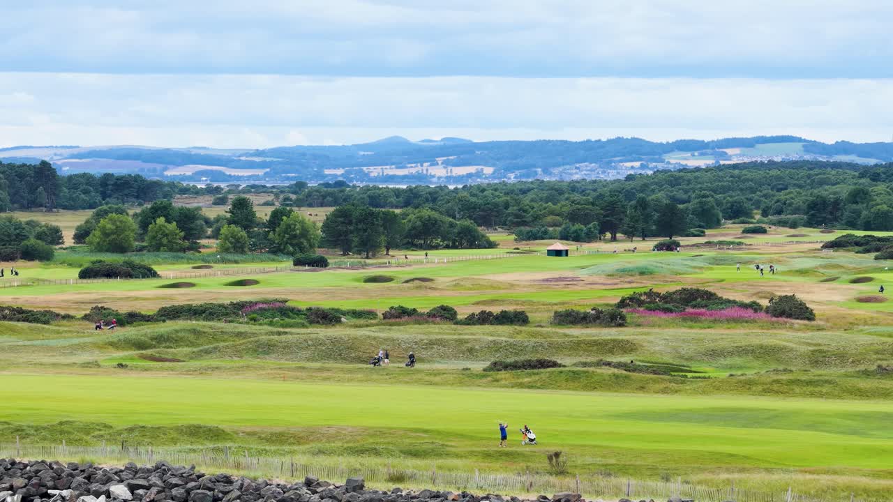 Wide shot of golfers walking on green fairway, rocky foreground, overcast sky, steady camera