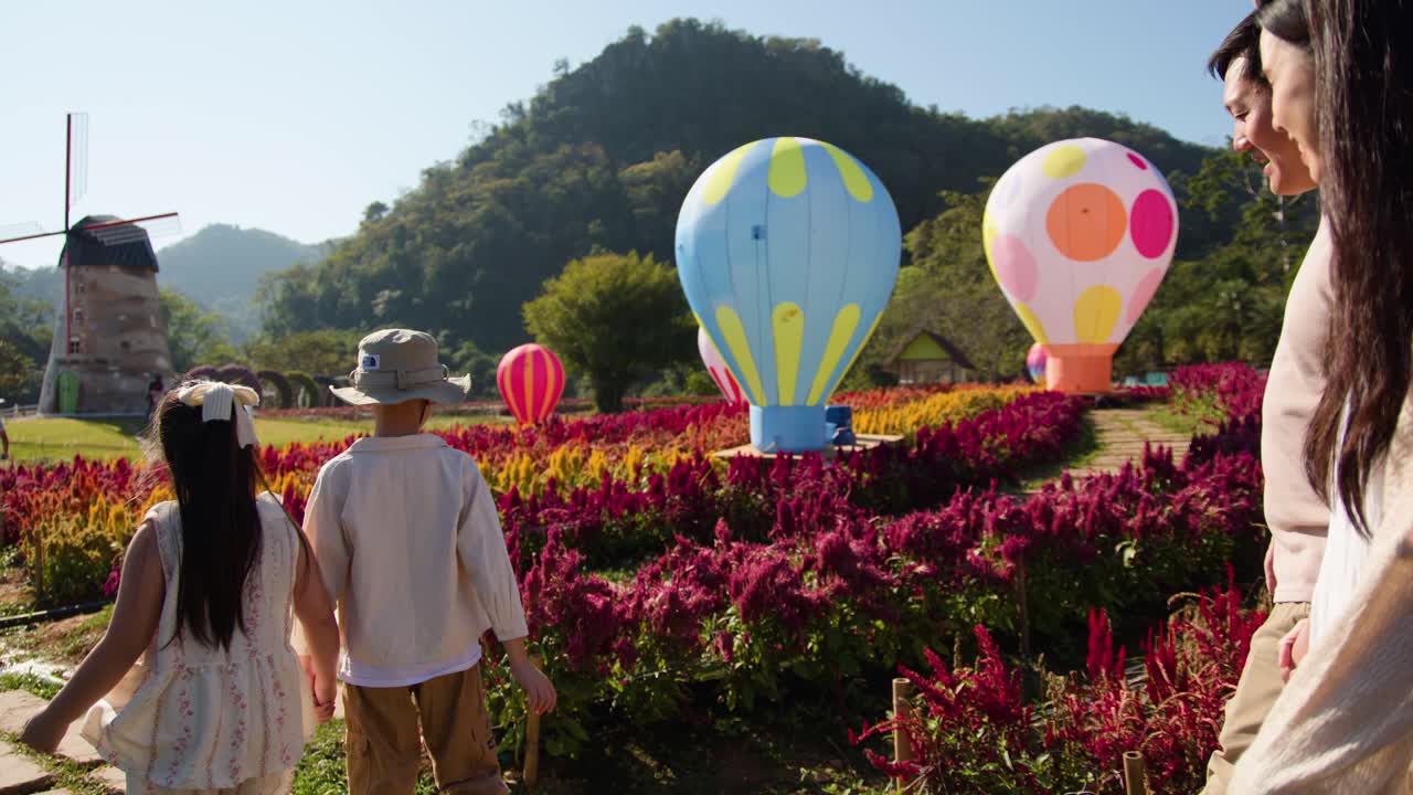 Parents and children stroll through vibrant flower garden with decorative hot air balloons, sunny daylight