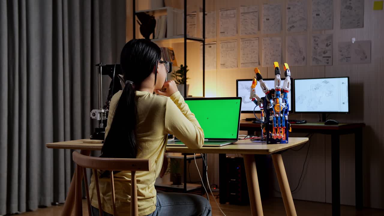 십대 아시아 소녀가 집에서 초록색 화면 모형을 가진 노트북에서 사이보그 손을 설계하는 동안 생각하는 뒷면