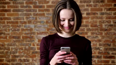 una joven hermosa está leyendo un mensaje en su teléfono inteligente, sonriendo, concepto de comunicación, fondo de ladrillo