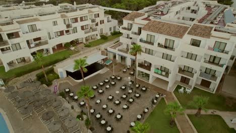 Vista-Aérea-De-Un-Complejo-Turístico-En-Mallorca,-España,-Que-Presenta-Un-Edificio-De-Alojamiento-De-Tres-Pisos-Con-Un-Patio-Delantero-Adornado-Con-Sillas-Y-Mesas.