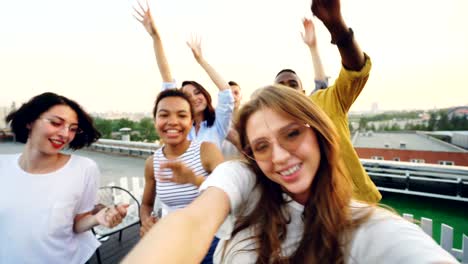 point of view shot of pretty young woman recording video of rooftop party with her friends dancing and looking at camera. entertainment, youth and music concept.