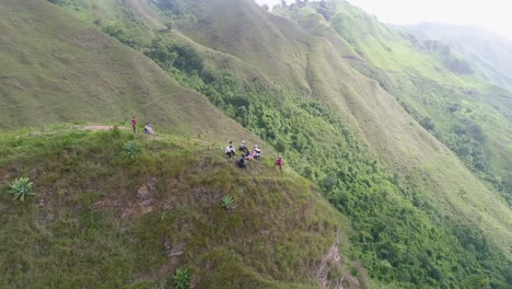 people-sitting-on-top-of-hill-enjoy-view-after-climbing,-drone-spiral-shot-over-green-mountains