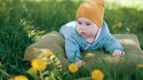 lovely baby in blue overall and orange cap lies on his belly. boy resting on the pillow in the green grass.