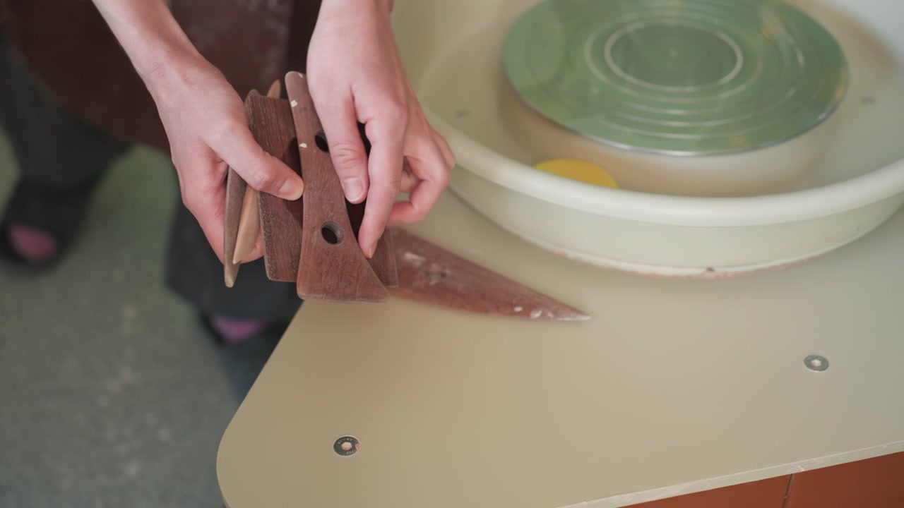 陶芸工房のろくろ道具がきちんと整理されている。使われていないろくろの横で、型紙、厚紙ガイド、削り具を置く手。スポンジとろくろ板が置かれた整頓されたスタジオテーブル、指導に適した整然とした配置