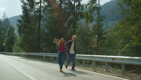 hitchhikers walking forest roadside in mountains. couple travelers holding sign.