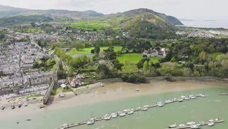 a large number of sailboats and motor vessels are moored at the long jetty in the fast flowing river off the english town of conwy among the green hills of wales on a cloudy day drone dolley tilt shot