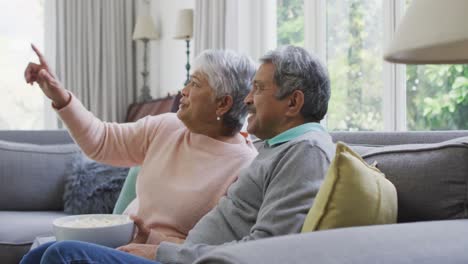 Happy-biracial-senior-couple-watching-tv-with-popcorn-and-having-fun-at-home