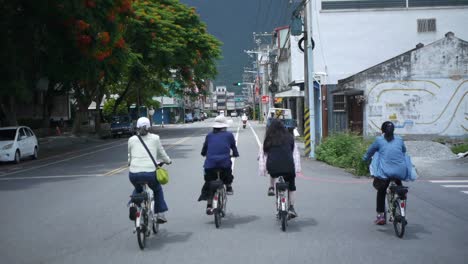 Group-of-4-woman-riding-bikes-through-countryside-town-in-Asia,-filmed-from-backside-following-riders
