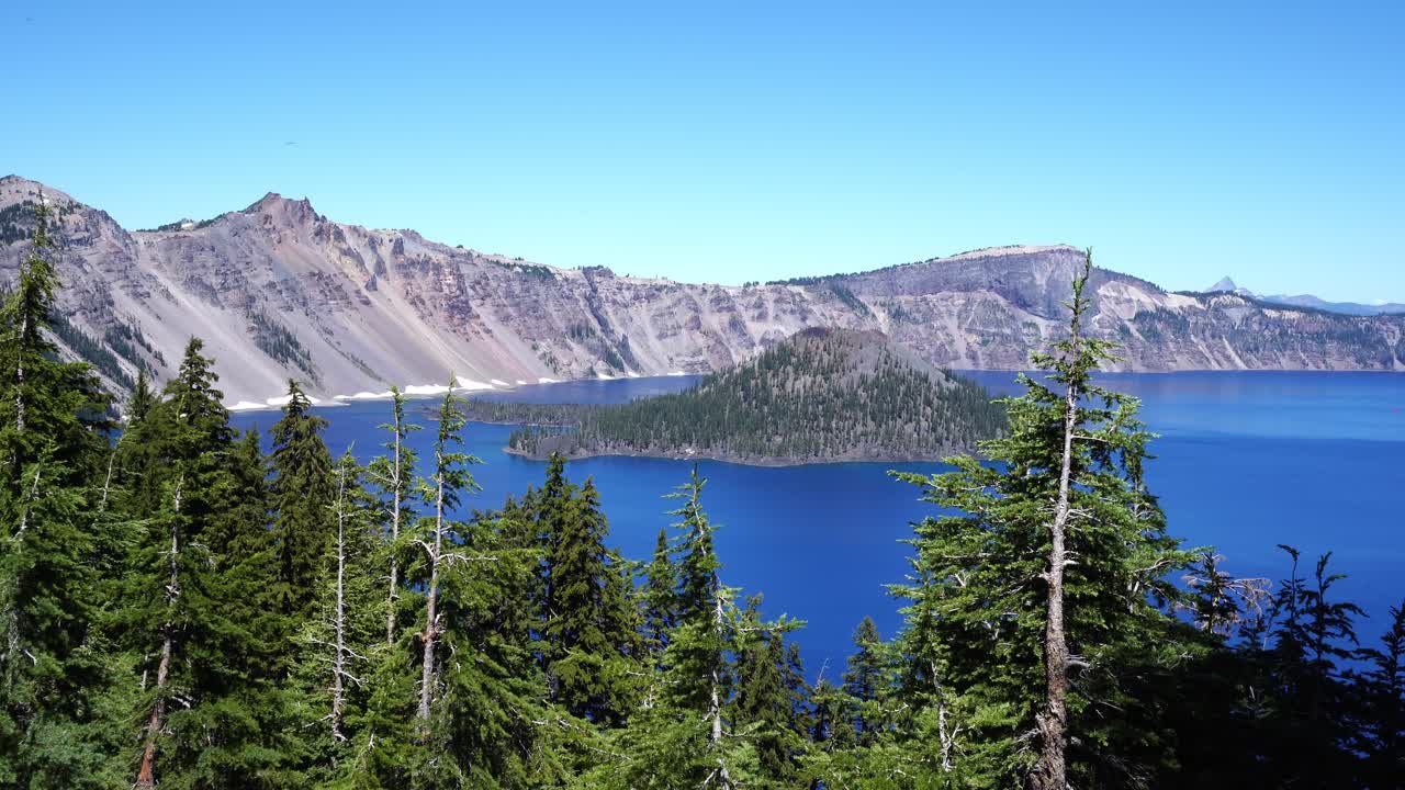 A vivid view of Crater Lake’s deep blue water and Wizard Island framed by tall evergreens under a clear summer sky in Oregon’s Crater Lake National Park