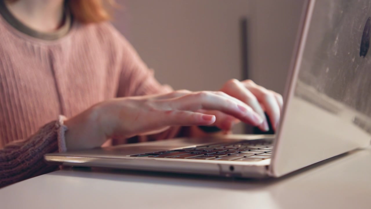 Primo piano da angolazione bassa mostra mani che digitano attivamente sulla tastiera di un laptop in un caldo spazio di lavoro domestico, con illuminazione laterale soffusa, profondità di campo ridotta, movimento visibile delle dita, avambracci e abbigliamento casual.