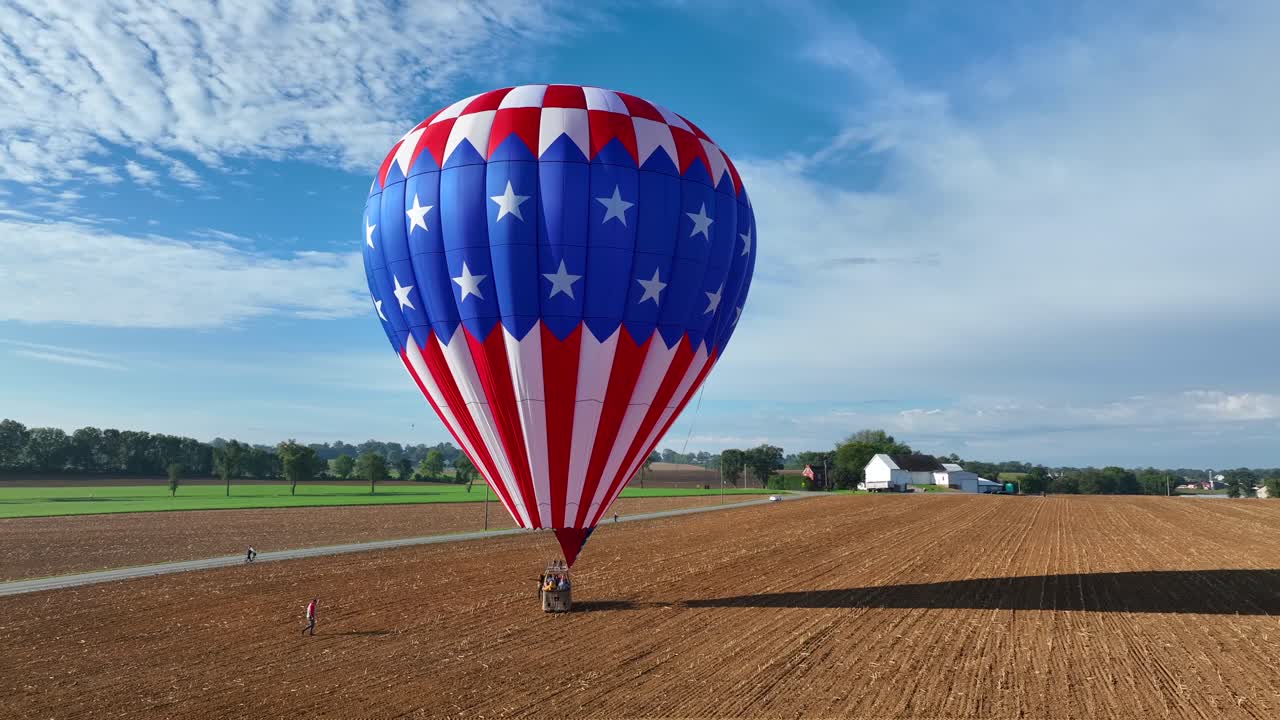 Aerial orbiting shot of american hot-air-balloon landing on agricultural field during sunlight