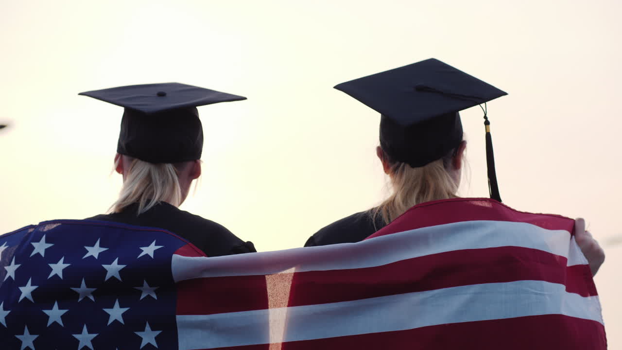 dos mujeres graduadas universitarias levantan las manos sobre los hombros de la bandera de estados unidos