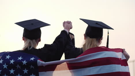 Two-Women-College-Graduates-Raise-Their-Hands-Up-On-The-Shoulders-Of-The-Usa-Flag