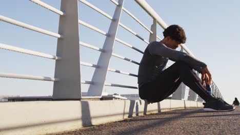 fit african american man exercising outdoors in city, resting sitting on footbridge