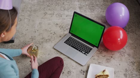 mujer sorprendida con sombrero de fiesta sosteniendo una caja de regalos mientras hace una videollamada en una computadora portátil en casa