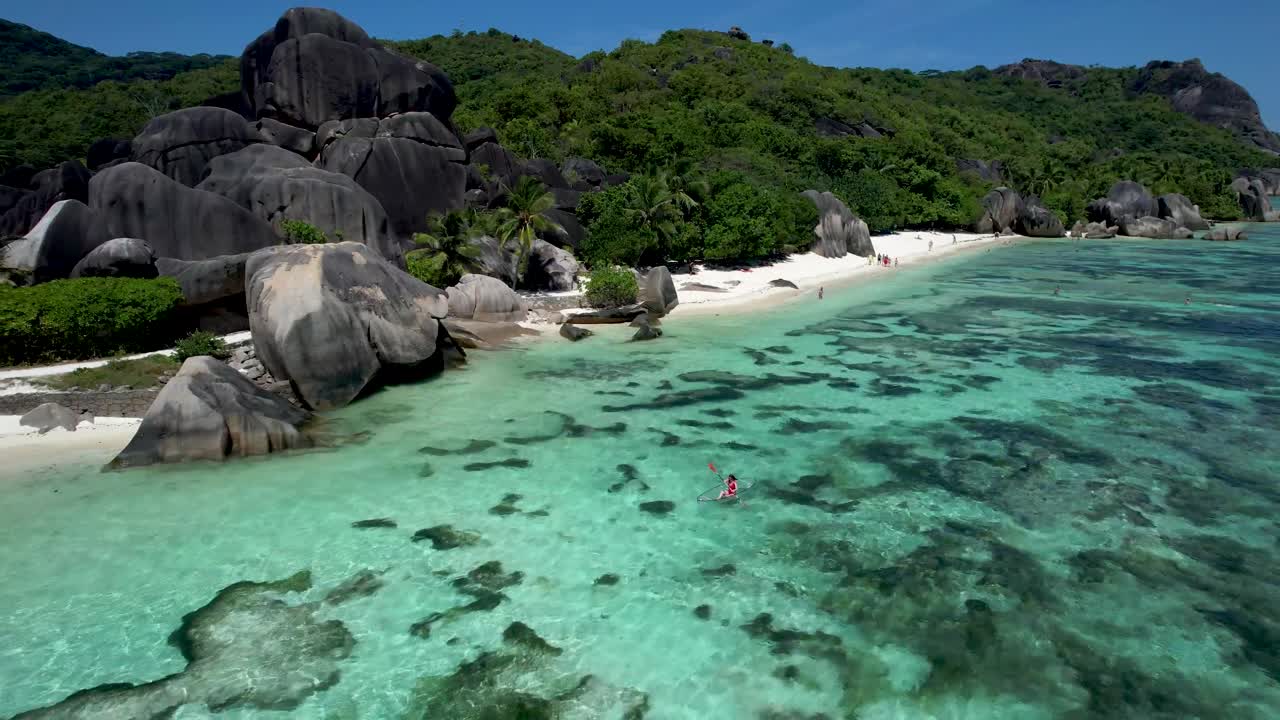 Donna che pagaia al largo della costa di un'iconica spiaggia delle Seychelles con massi e palme.