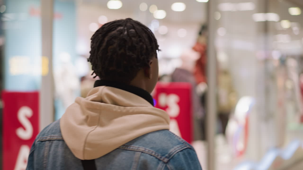 young black man facing storefront in shopping mall wearing denim jacket and hoodie, viewing window displays of mannequins and sale items, indoor retail with modern lighting and glass architecture