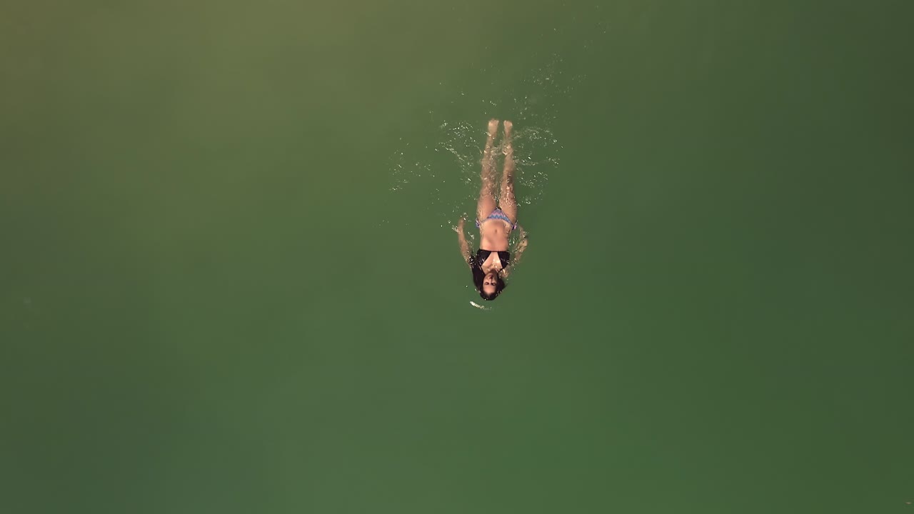 воздушный снимок сверху вниз слежение за женщиной в бикини, расслабленно плавающей в красивой зеленой поверхности океанской воды