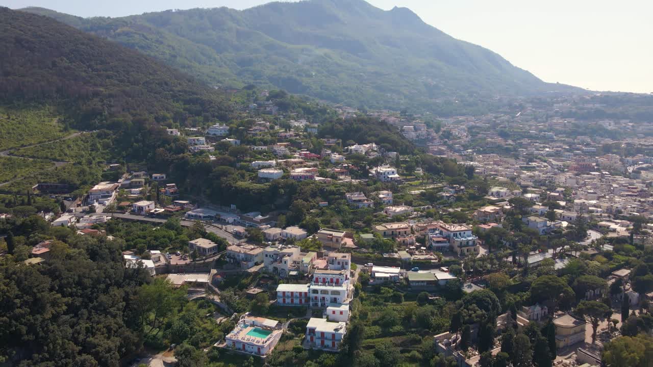 이탈리아 이스키아 열대 섬의 해안선에 있는 스파를 드러내며 아래로 천천히 패닝하는 마을과 멀리 있는 화산의 광각 드론 샷