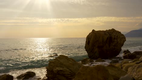 seascape at twilight with sun rays flares on sea surface seen through rocky beach in mediterranean