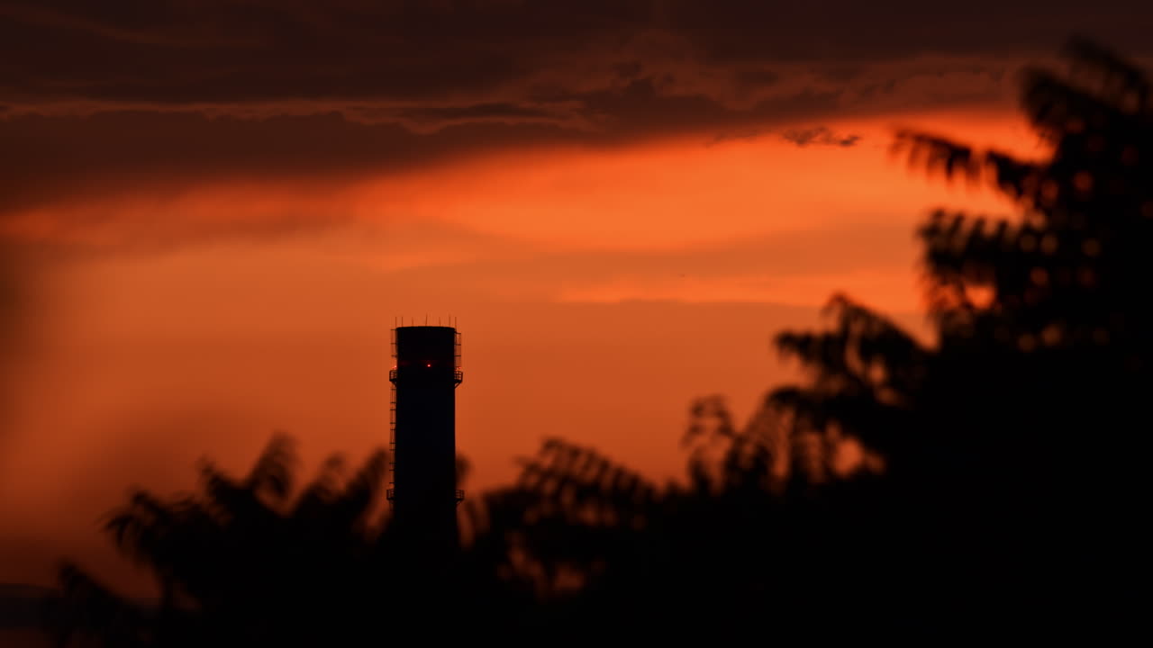 An industrial chimney stands against a colorful sunset. The sky transitions from orange to dark hues