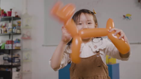 little girl playing with a long balloon in the shape of a dog while looking at camera in classroom in a montessori school