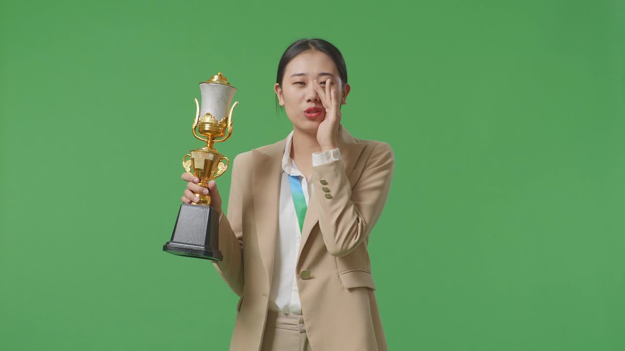 금메달을  슈트를 입은 아시아 사업가 여성이 금 트로피를 들고 스튜디오에서 녹색 스크린 배경에 입을 고 긍정적으로 소리치고 있습니다.