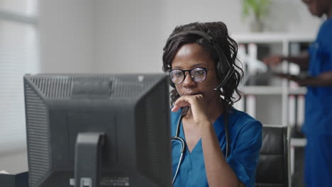 a black woman doctor wearing headphones sits at a table with a computer and takes calls from patients looks at their medical records and enters them into the clinic schedule