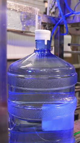 big full bottles of clean water move by the conveyor belt. machine is lifting a bottle and worker covers it in plastic bag. vertical video