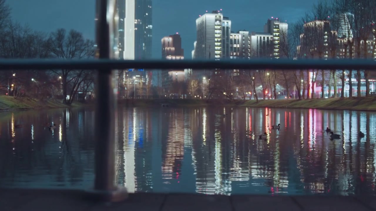 Night city skyline reflected in water, empty boardwalk and metal railing foreground, highrise lights mirrored on calm river, cool blue tones and soft bokeh, tranquil urban atmosphere for architecture
