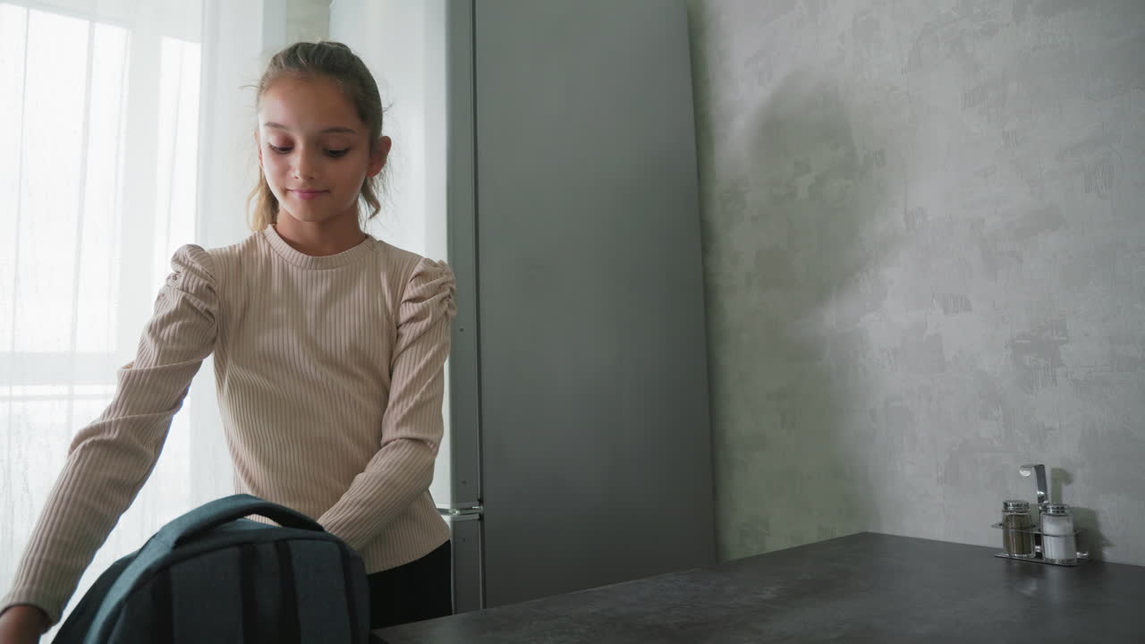 Girl in casual beige blouse packing water bottle into backpack on kitchen counter with lunch container nearby, preparing for school in soft morning light next to refrigerator and window curtains