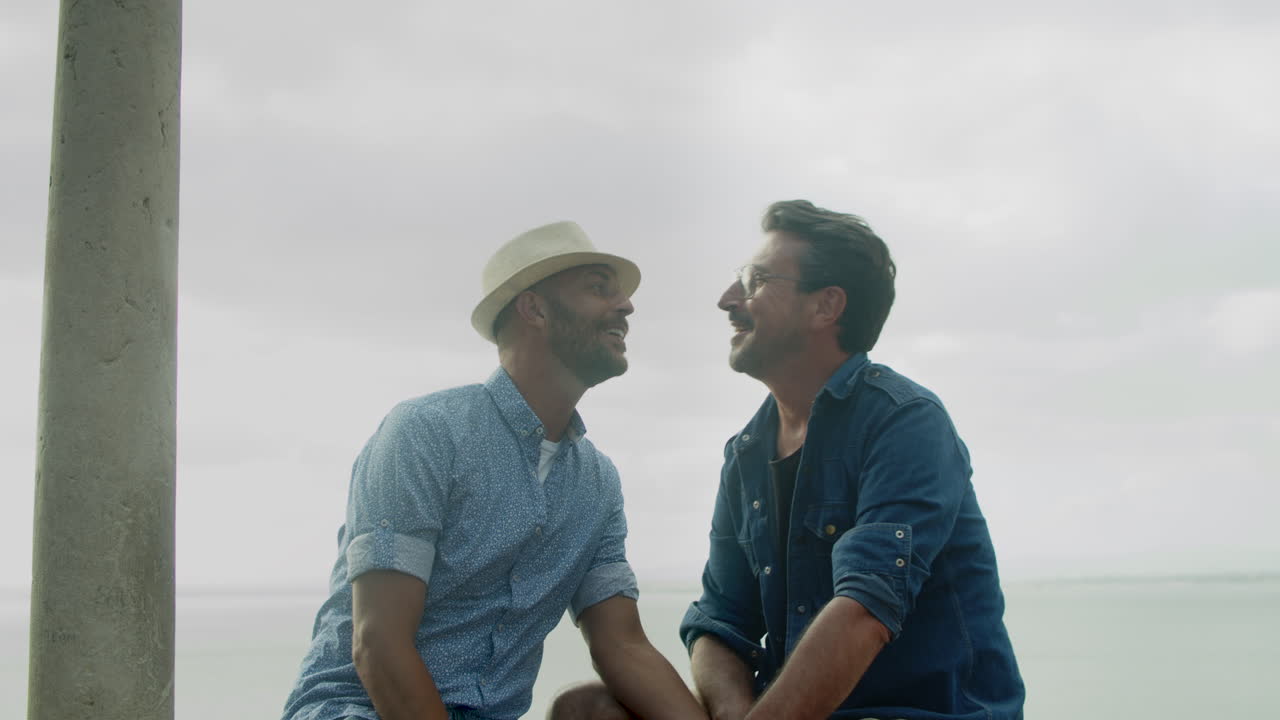 Vista frontale di una coppia gay seduta con il mare sullo sfondo e che si bacia