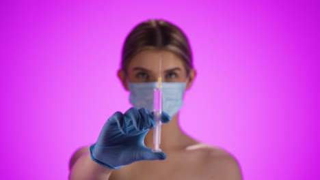 Medium-shot-of-a-young-pretty-woman-wearing-a-ffp2-mask-while-holding-a-syringe-with-purple-liquid-into-the-camera-to-vaccinate-against-diseases-or-corona-virus-in-front-of-purple-background