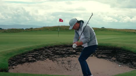 lefty golfer hits sand shot out of ireland pot bunker onto green in slow motion, ireland links golf on beautiful cloudy day in 4k