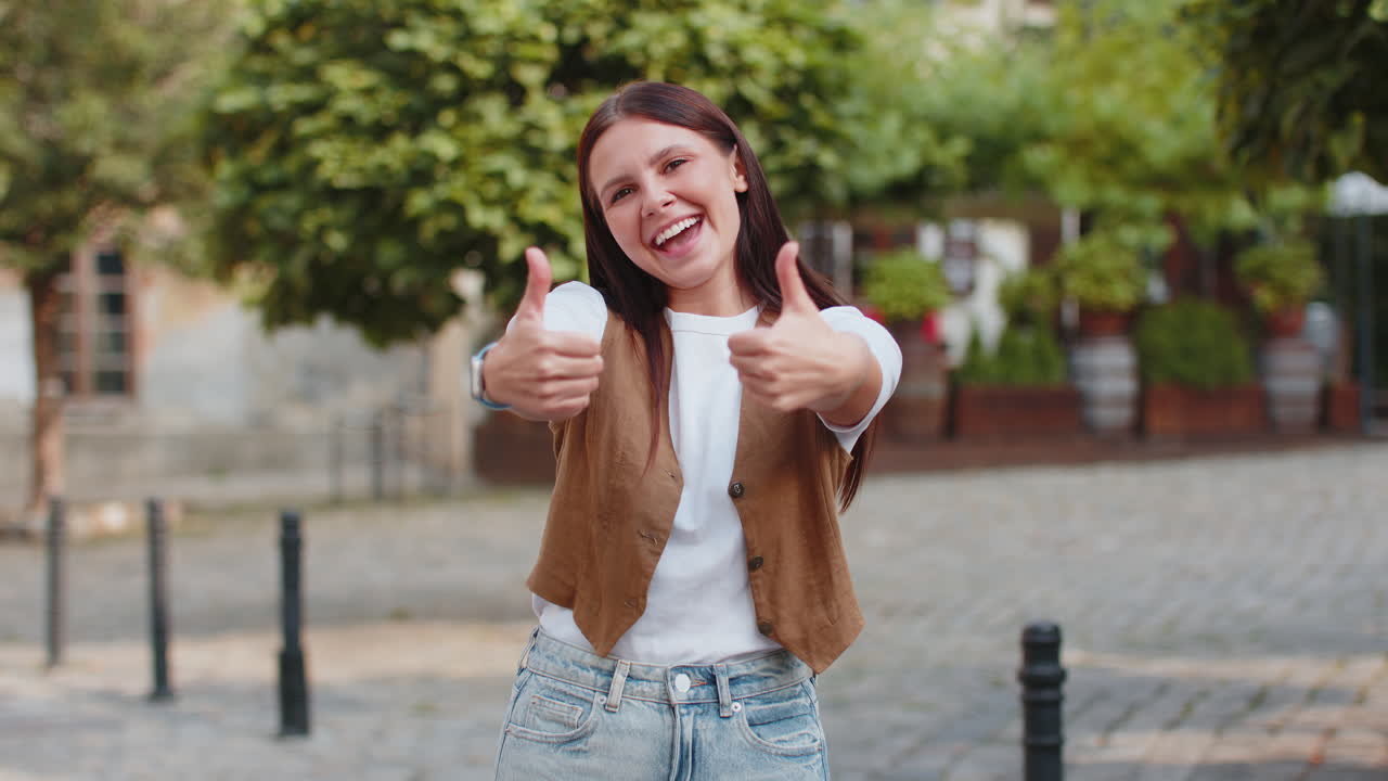 Cheerful young caucasian woman showing double thumbs up gives positive reply standing on city street