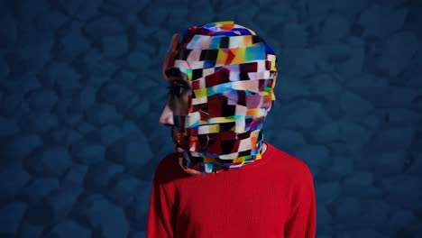 person with colorful geometric mask standing against an orange and blue striped background, creating a surreal and artistic portrait exploring themes of identity, disguise, and modern art