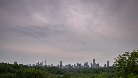 vertically panning down timelapse of the city skyline in toronto canada during sunset with a cloudy pink and blue sky as traffic passes by on a nearby highway surrounded by a forest of trees.