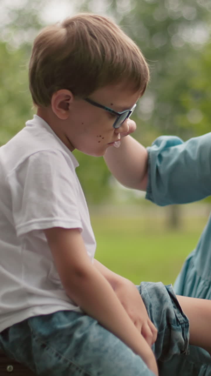 una madre se arrodilla para limpiar la herida de su hijo mientras se sientan a lo largo de un camino asfalto, el niño sostiene sus pantalones, mirando triste, mientras su bicicleta caída yace cerca, la madre suavemente limpia sus lágrimas