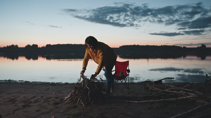 Campeggio in riva al lago al crepuscolo. Una donna sistema la legna e getta rami su una catasta vicino all'acqua calma, con una sedia pieghevole rossa sullo sfondo. Movimento e intento mentre il campeggio viene preparato all'imbrunire, sotto un cielo riflesso e nella quiete.