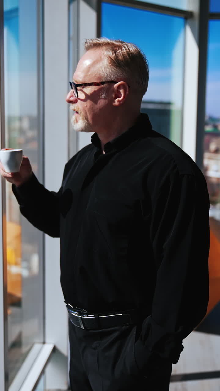 Confident senior man wearing black clothes stands at the panoramic window. Man looks at city panorama, drinks coffee and turns to smile to camera. Vertical video.