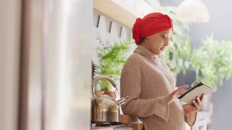 video of smiling biracial woman in hijab using tablet standing in kitchen at home