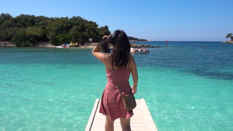 Girl-in-red-summer-dress-walking-on-pier-out-towards-turquoise-ocean-water-in-slow-motion