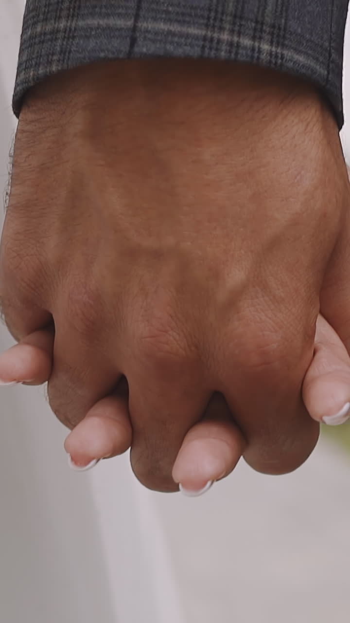 Bride and groom in festive outfit walk joining hands with interlaced fingers at wedding ceremony closeup slow motion. Newlywed young man and woman relationship