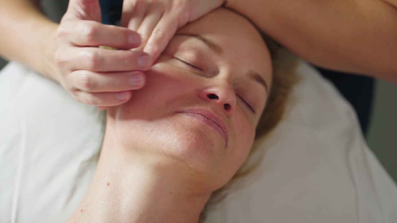High angle close up relaxed client on massage bed under ring light as practitioner gently tilts head sideways and massages cheek and jawline with smooth strokes during calming facial session