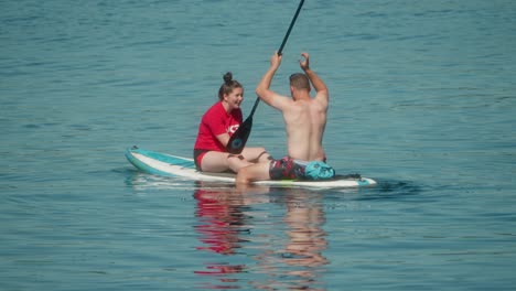 a young couple laughs and paddles together on a sunny day, sharing a playful moment while seated on a stand-up paddleboard in calm, clear water